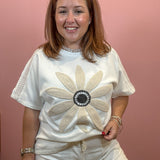 A smiling woman wearing a white short-sleeve t-shirt with a large beige and black beaded flower applique on the front, paired with white shorts, set against a solid dusty pink wall.