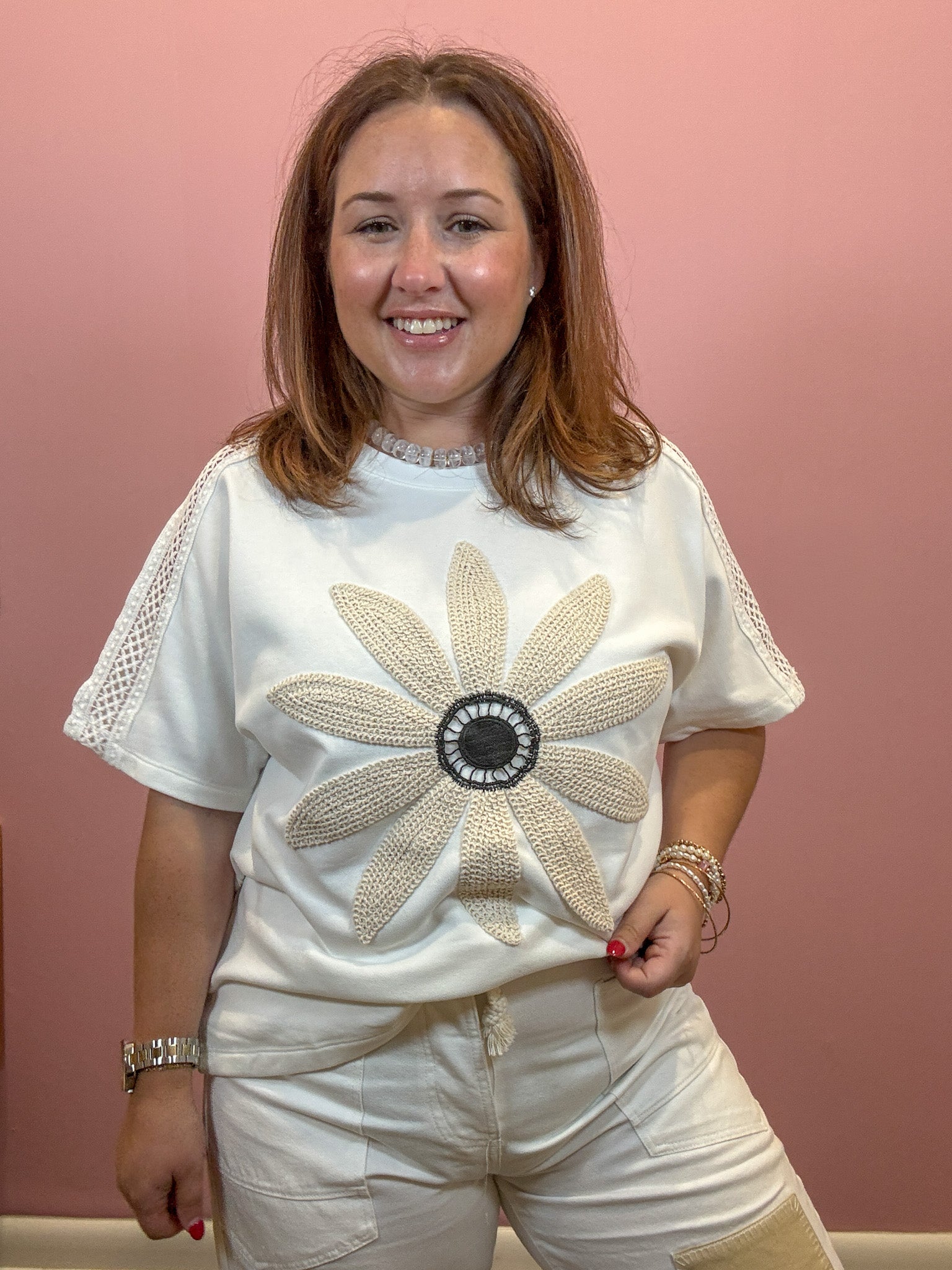 A smiling woman wearing a white short-sleeve t-shirt with a large beige and black beaded flower applique on the front, paired with white shorts, set against a solid dusty pink wall.