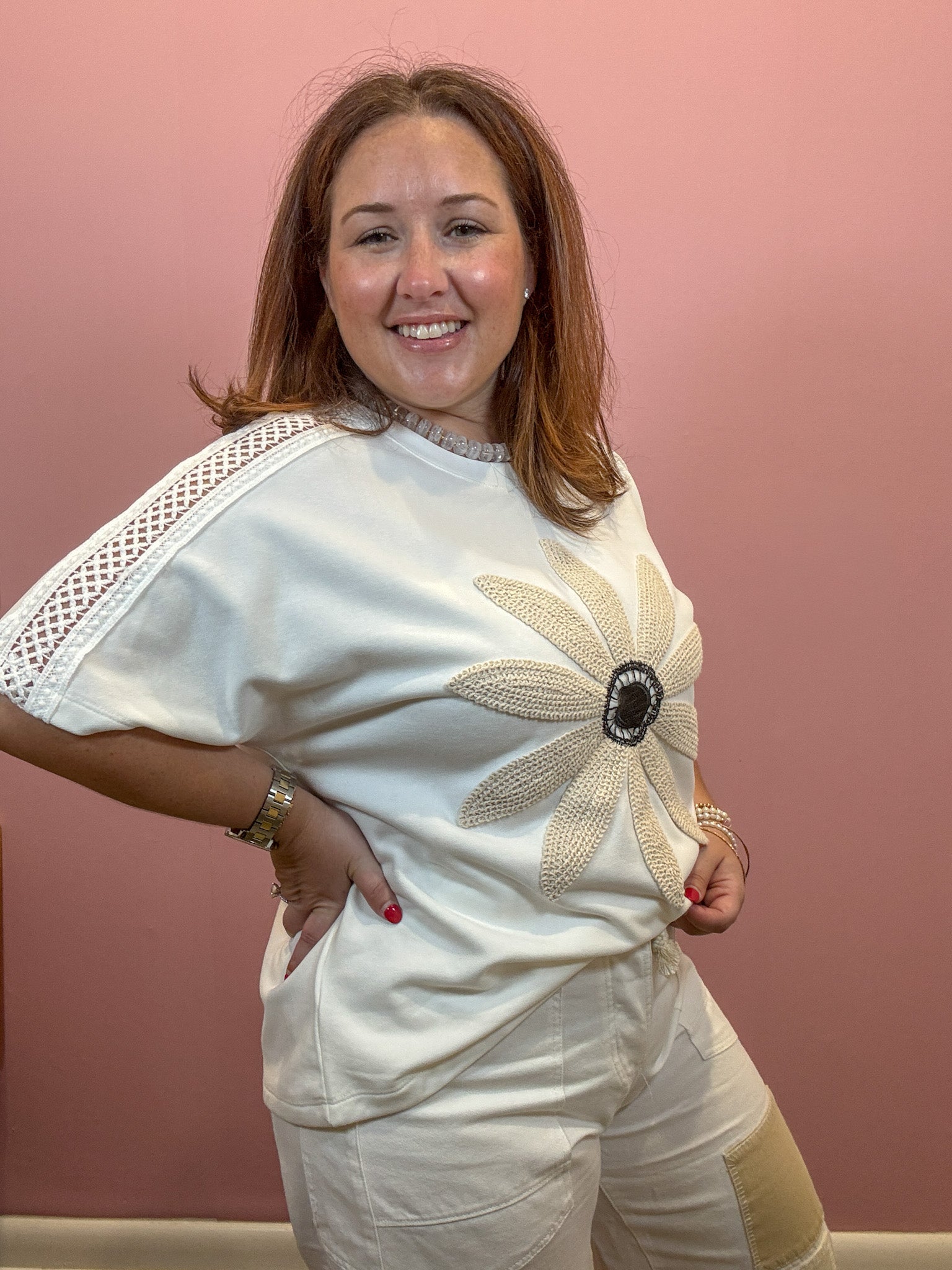 A smiling woman wearing a cream-colored short-sleeve top with a large floral applique on the front and matching cream pants, posing against a solid dusty pink wall.