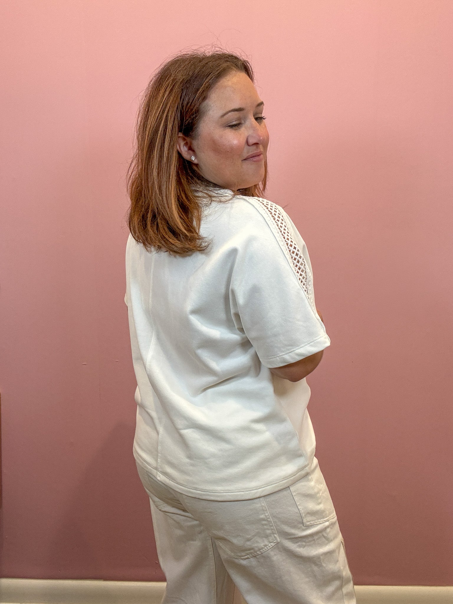 A woman with shoulder-length brown hair is shown from the back wearing a white short-sleeve top with a textured crochet detail on the shoulder and white pants, standing against a solid pink wall.