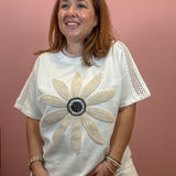 A woman wearing a white short-sleeve t-shirt with a large beige beaded flower design on the front and beige crochet lace sleeves, paired with beige pants, standing against a dusty pink wall.