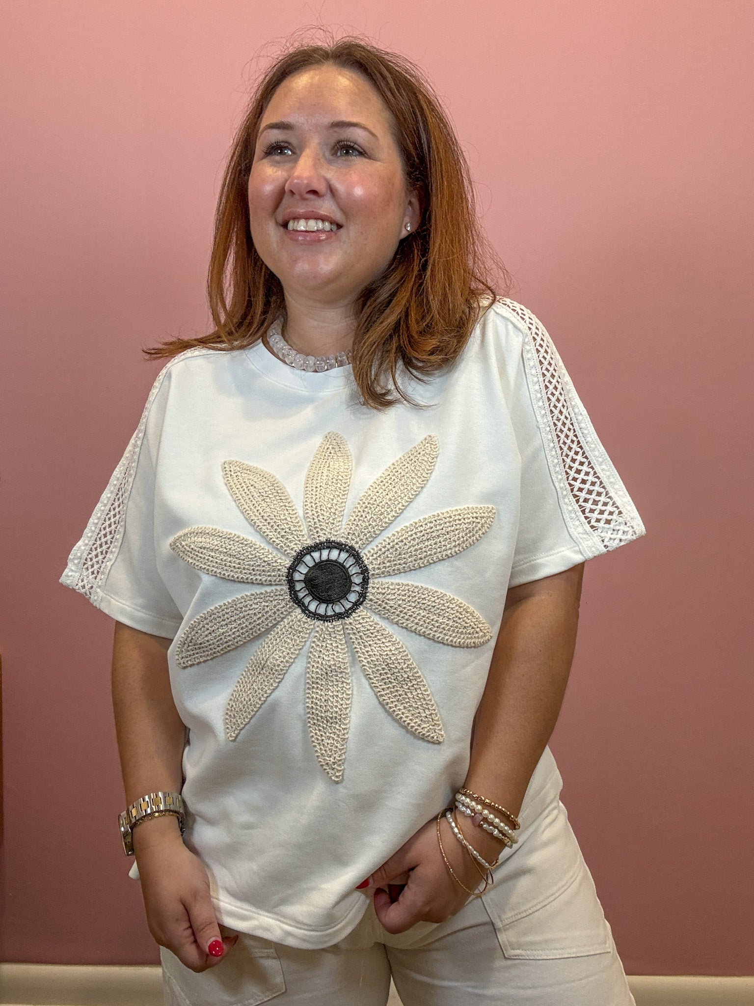 A woman wearing a white short-sleeve t-shirt with a large beige beaded flower design on the front and beige crochet lace sleeves, paired with beige pants, standing against a dusty pink wall.
