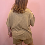 A woman viewed from behind wearing a matching beige short-sleeve top and shorts set against a solid pink wall.