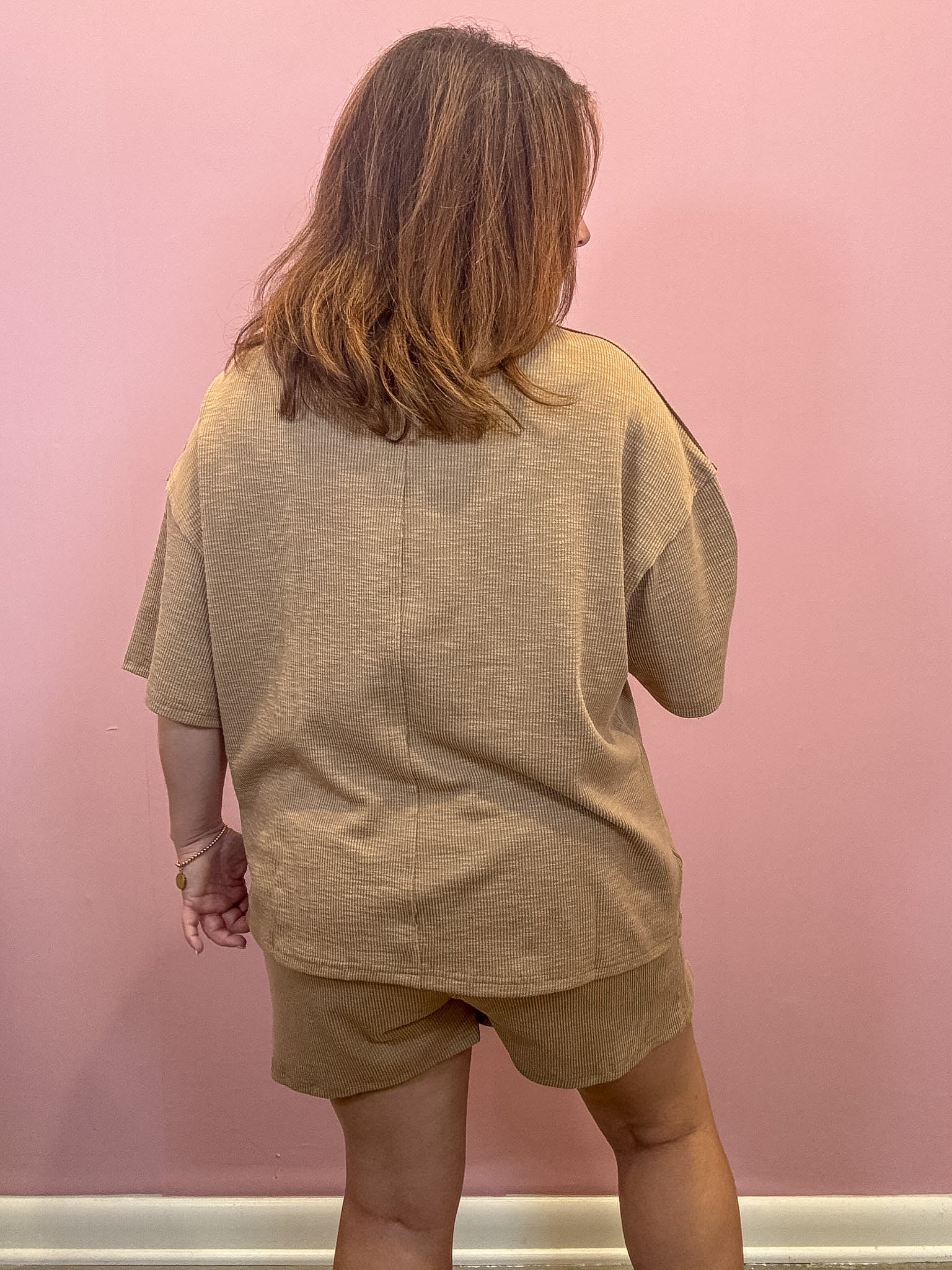 A woman viewed from behind wearing a matching beige short-sleeve top and shorts set against a solid pink wall.