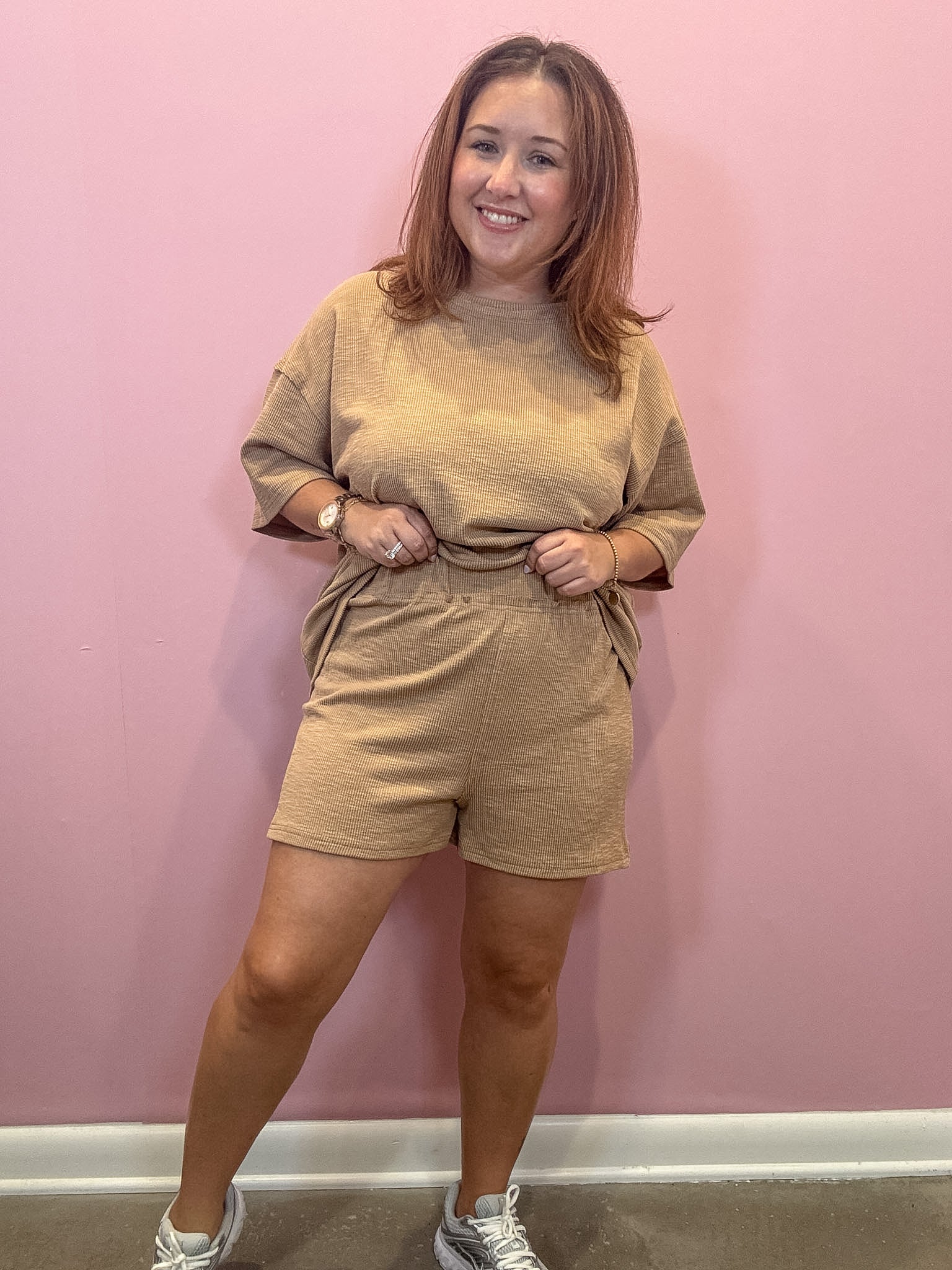 A woman modeling a matching beige lounge set consisting of a short-sleeve crewneck top and matching shorts, standing against a solid pink wall.