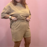 A woman modeling a matching beige lounge set consisting of a short-sleeve crewneck top and coordinating shorts, standing against a pink wall.