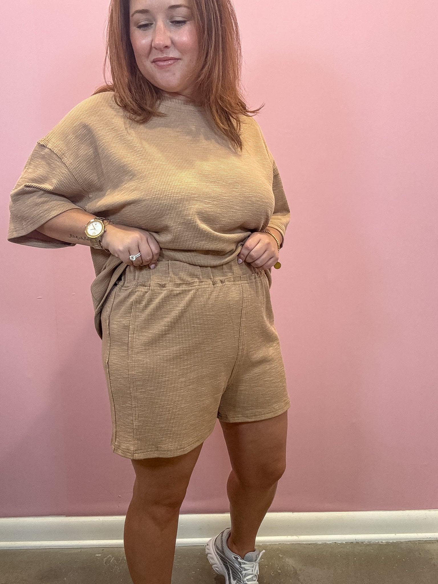 A woman modeling a matching beige lounge set consisting of a short-sleeve crewneck top and coordinating shorts, standing against a pink wall.