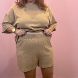 A woman modeling a matching beige lounge set consisting of a short-sleeve crewneck sweatshirt and high-waisted shorts, posed against a solid pink wall.