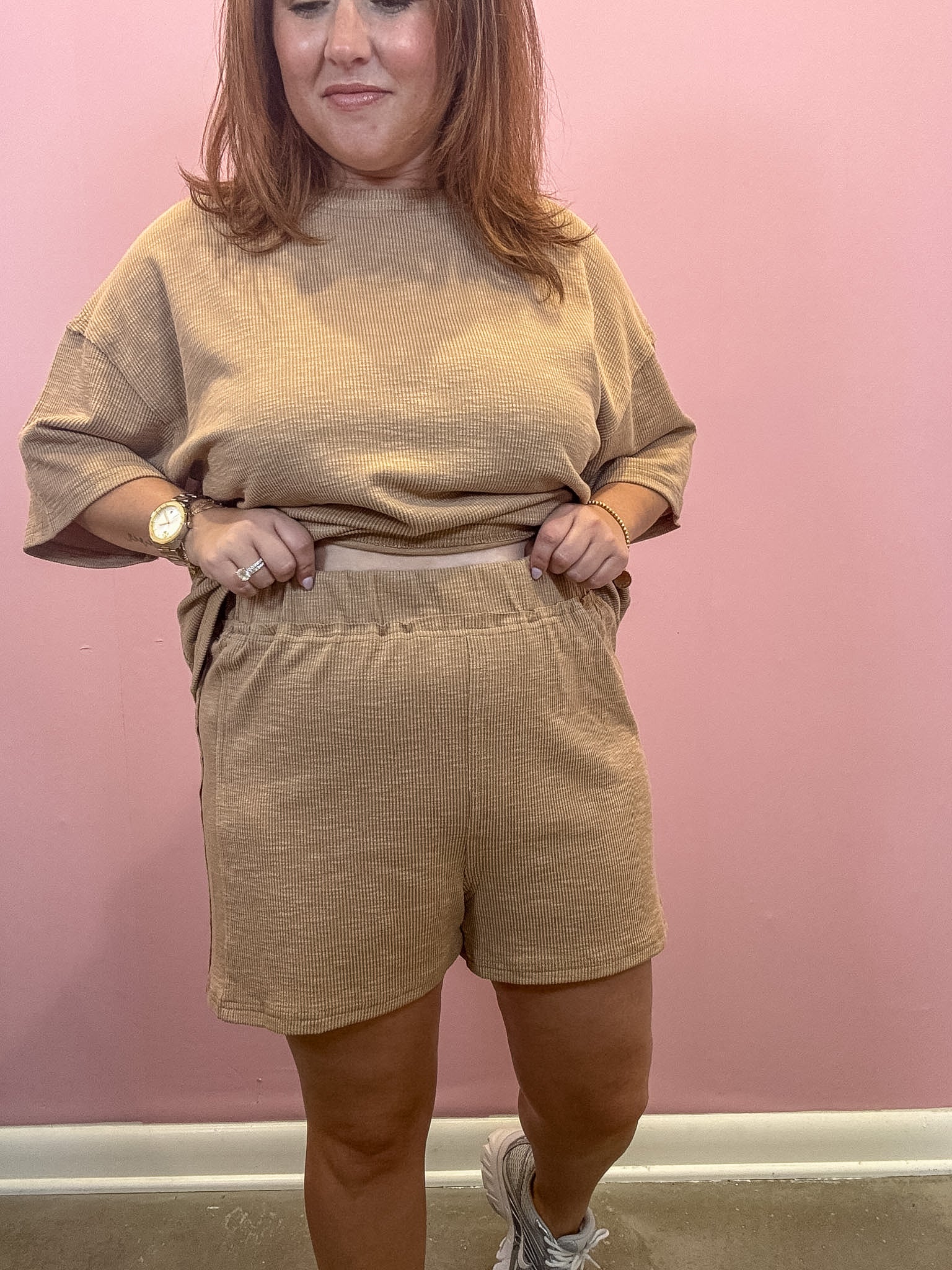 A woman modeling a matching beige lounge set consisting of a short-sleeve crewneck sweatshirt and high-waisted shorts, posed against a solid pink wall.