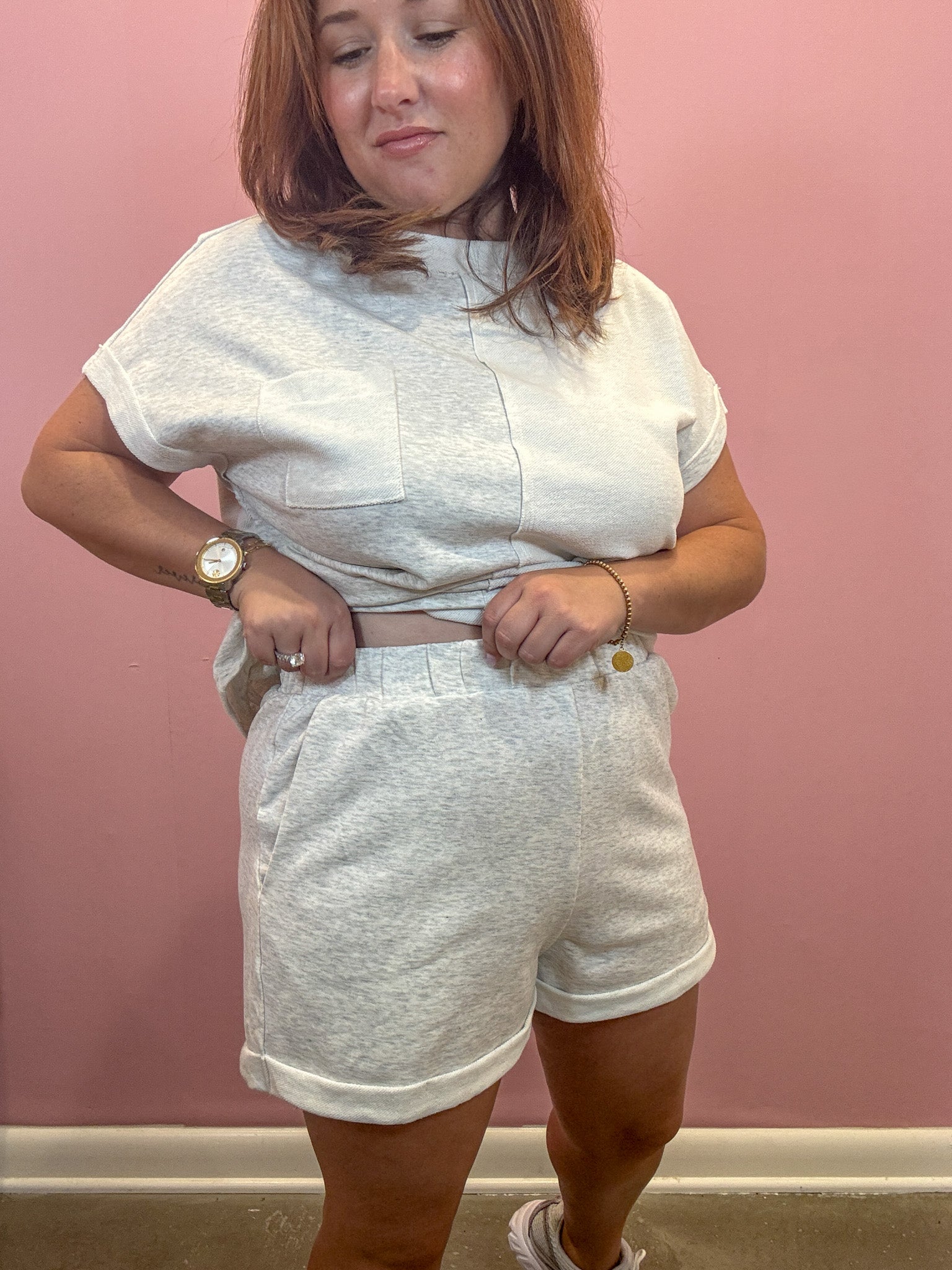 A woman modeling a matching light gray heather lounge set consisting of a short-sleeve cropped t-shirt and shorts, standing against a pink wall.