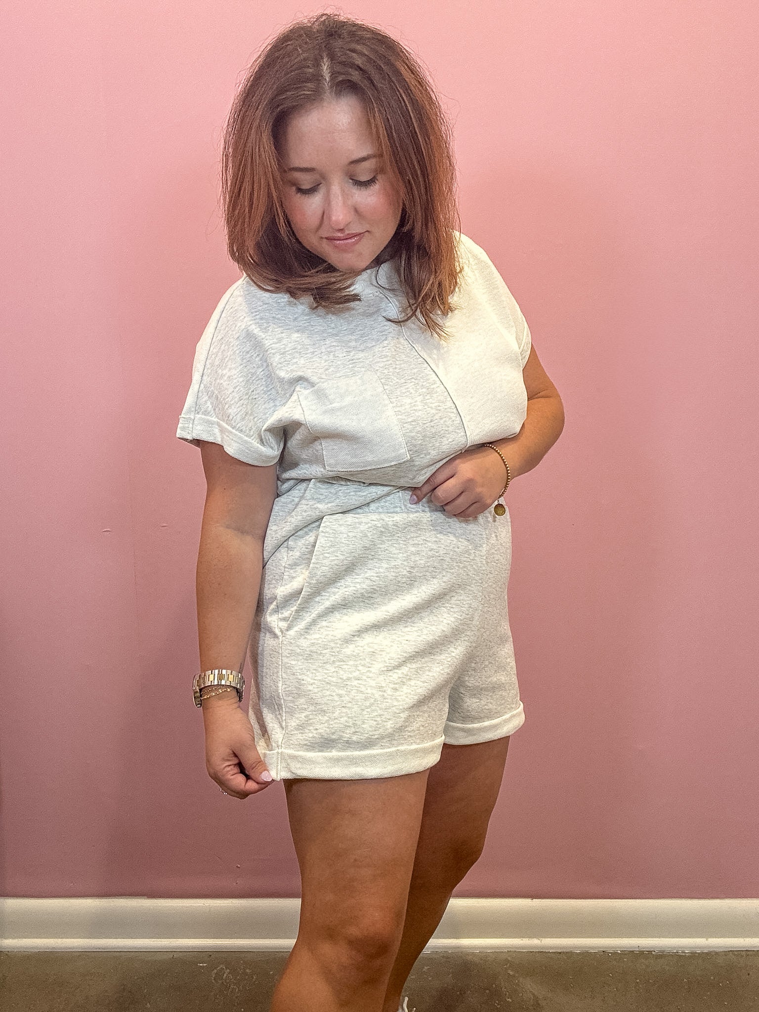 A woman modeling a light gray heathered short-sleeve t-shirt and matching shorts set against a solid pink wall.