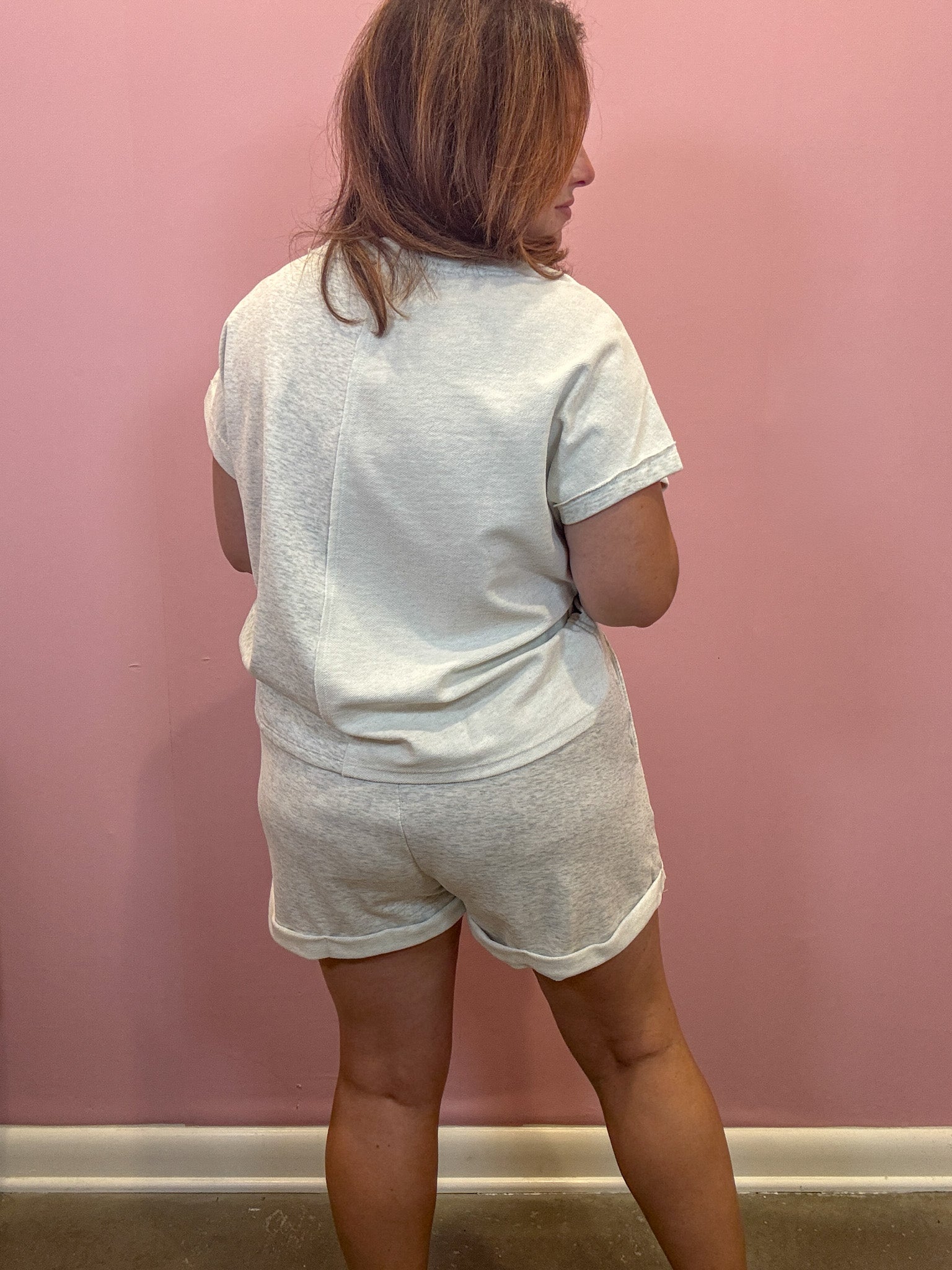 A woman viewed from behind wearing a light gray heathered t-shirt and matching shorts against a pink wall.