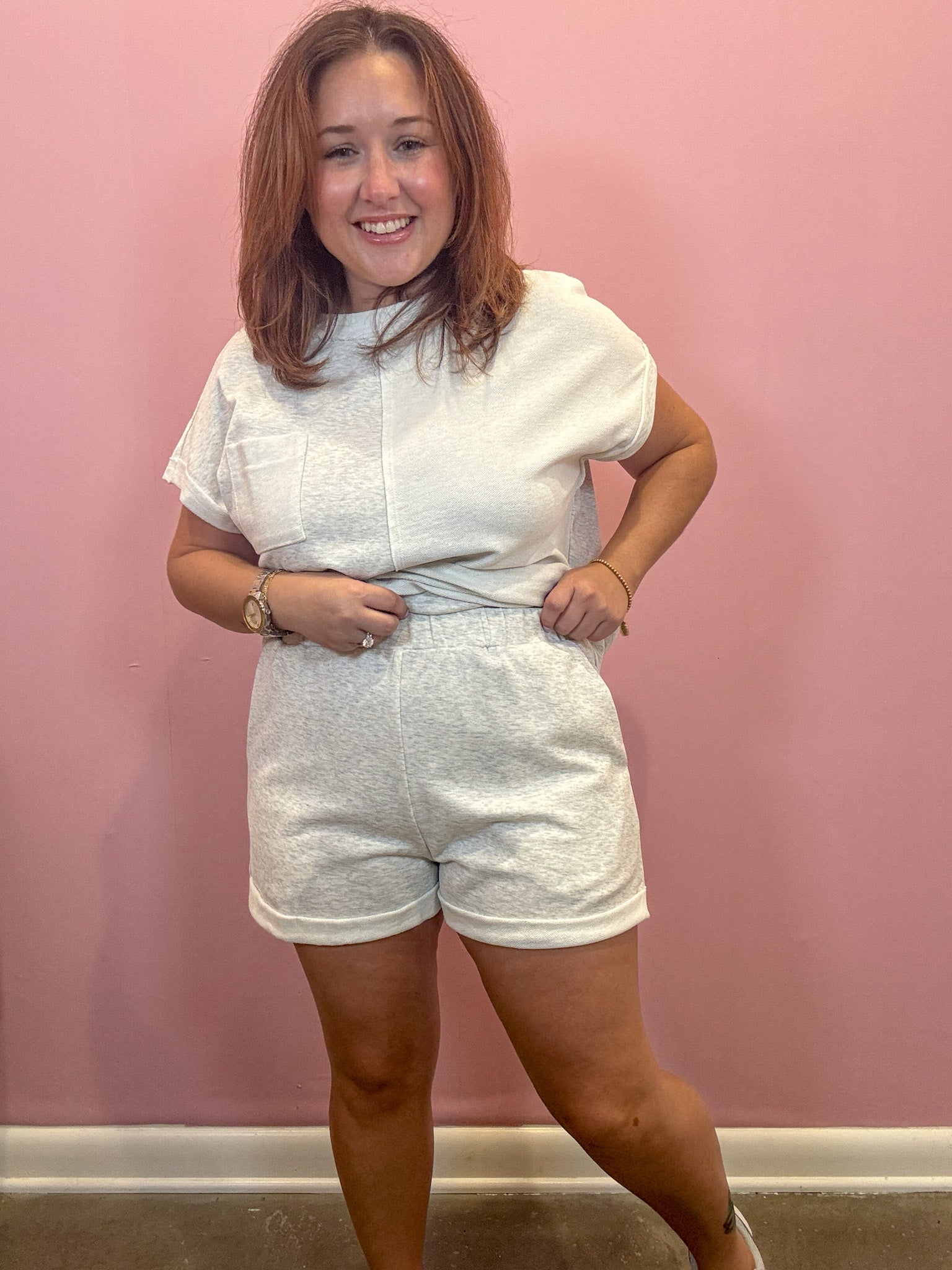A woman modeling a matching white two-piece lounge set consisting of a short-sleeve top and textured shorts, standing against a pink wall.