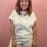A woman wearing a white textured short-sleeve top and matching shorts, standing against a solid pink wall.