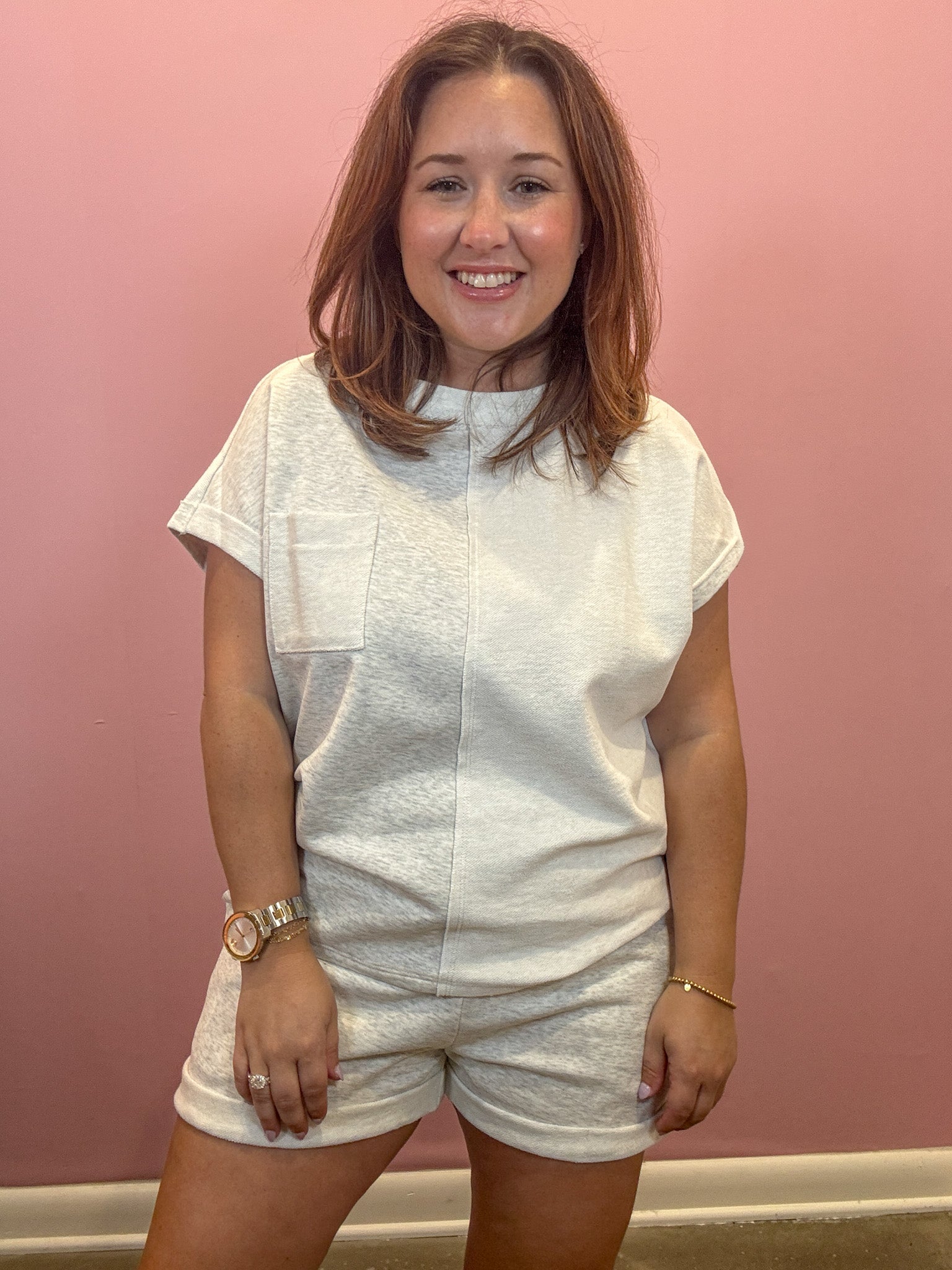 A woman wearing a white textured short-sleeve top and matching shorts, standing against a solid pink wall.