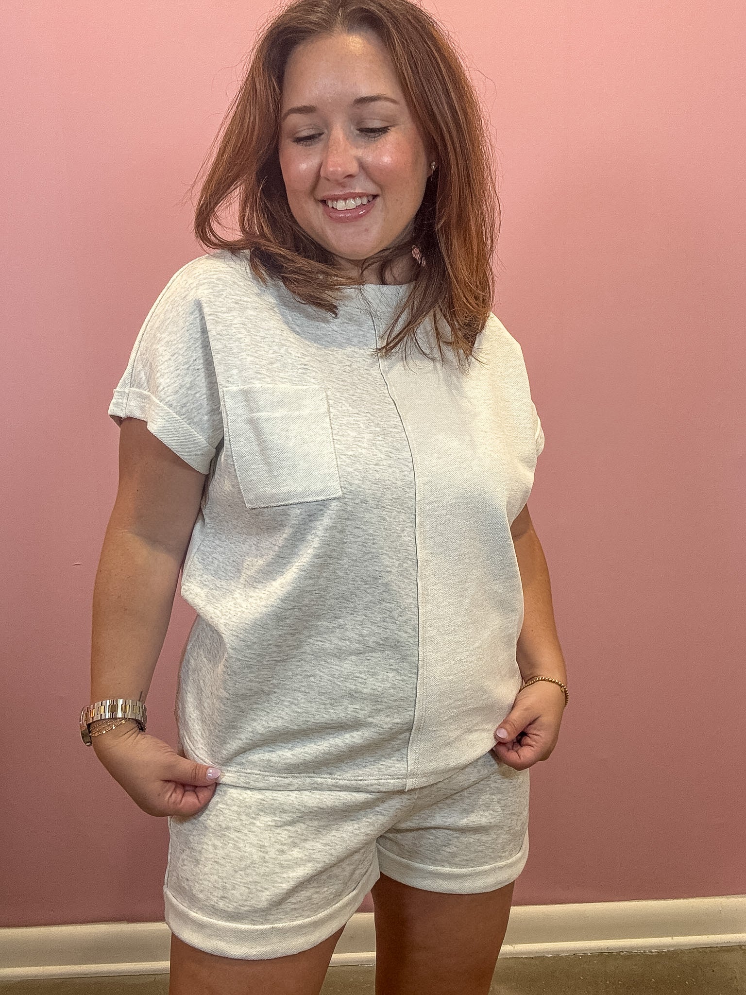 A woman wearing a light heather gray short-sleeve t-shirt and matching shorts, standing against a solid pink wall.