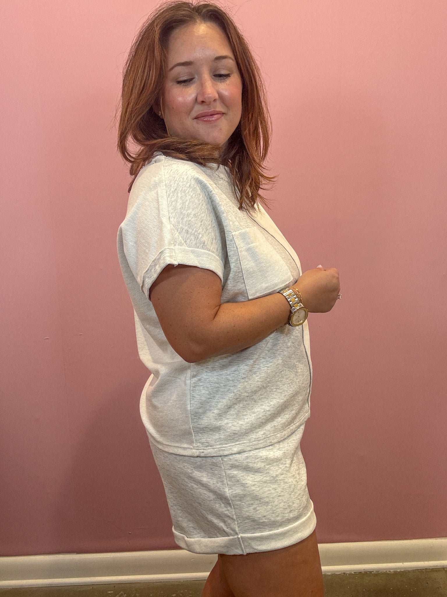 A woman wearing a light gray heathered short-sleeve t-shirt and matching shorts, standing against a solid pink wall.