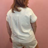 A woman viewed from behind wearing a white short-sleeve t-shirt and matching white shorts against a pink wall.