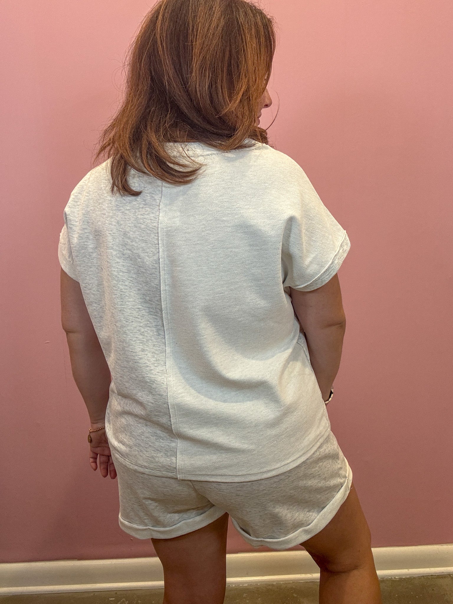 A woman viewed from behind wearing a white short-sleeve t-shirt and matching white shorts against a pink wall.