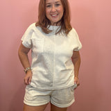 A woman modeling a matching white two-piece lounge set consisting of a short-sleeve top with a chest pocket and coordinating shorts, standing against a solid pink wall.