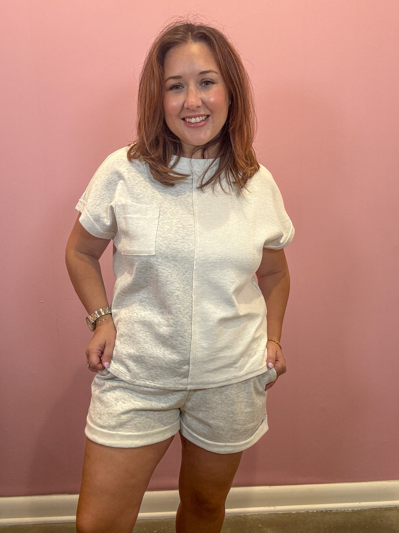 A woman modeling a matching white two-piece lounge set consisting of a short-sleeve top with a chest pocket and coordinating shorts, standing against a solid pink wall.