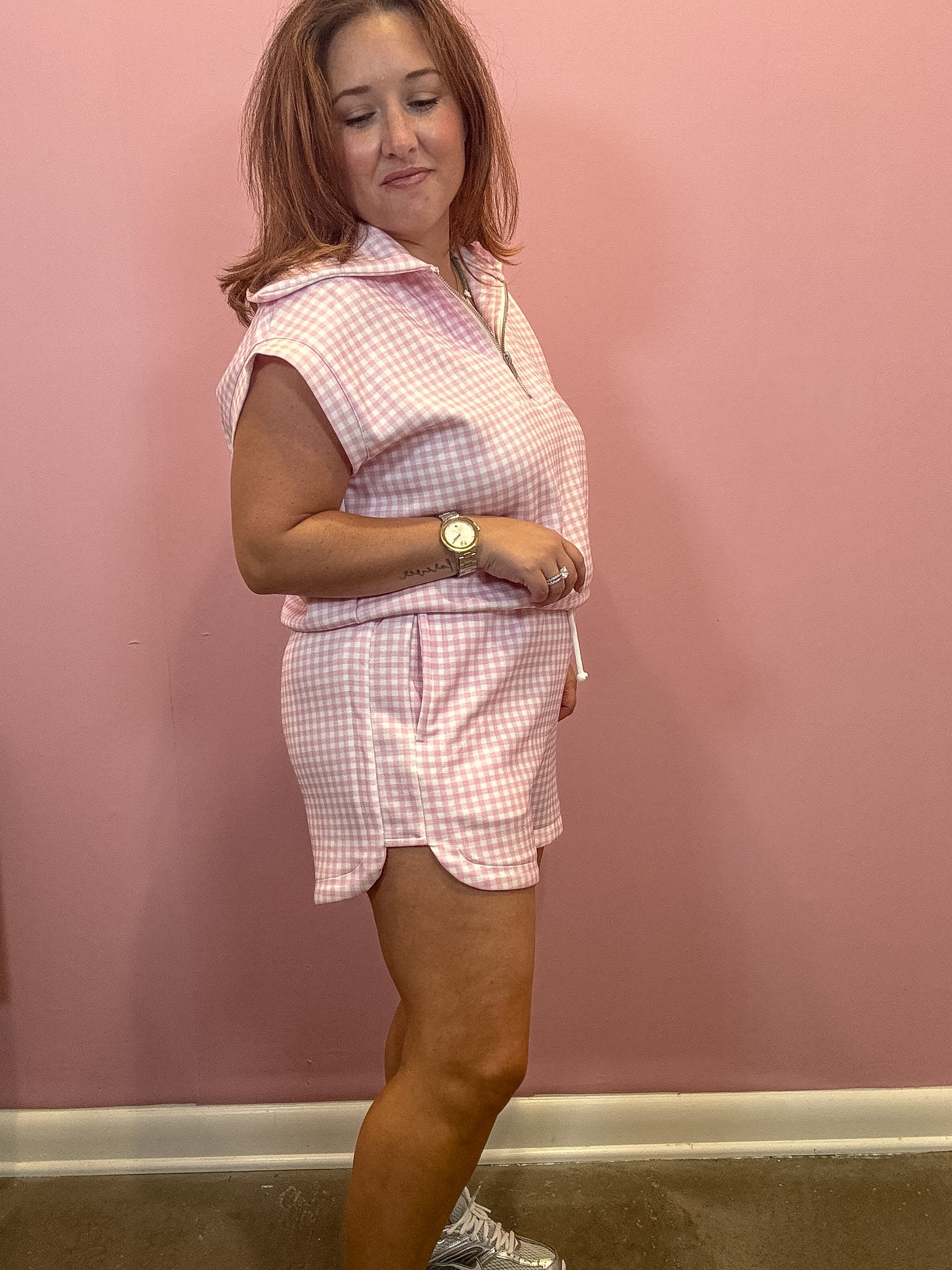 A woman modeling a pink and white gingham matching set consisting of a sleeveless collared top and shorts, standing against a solid pink wall.
