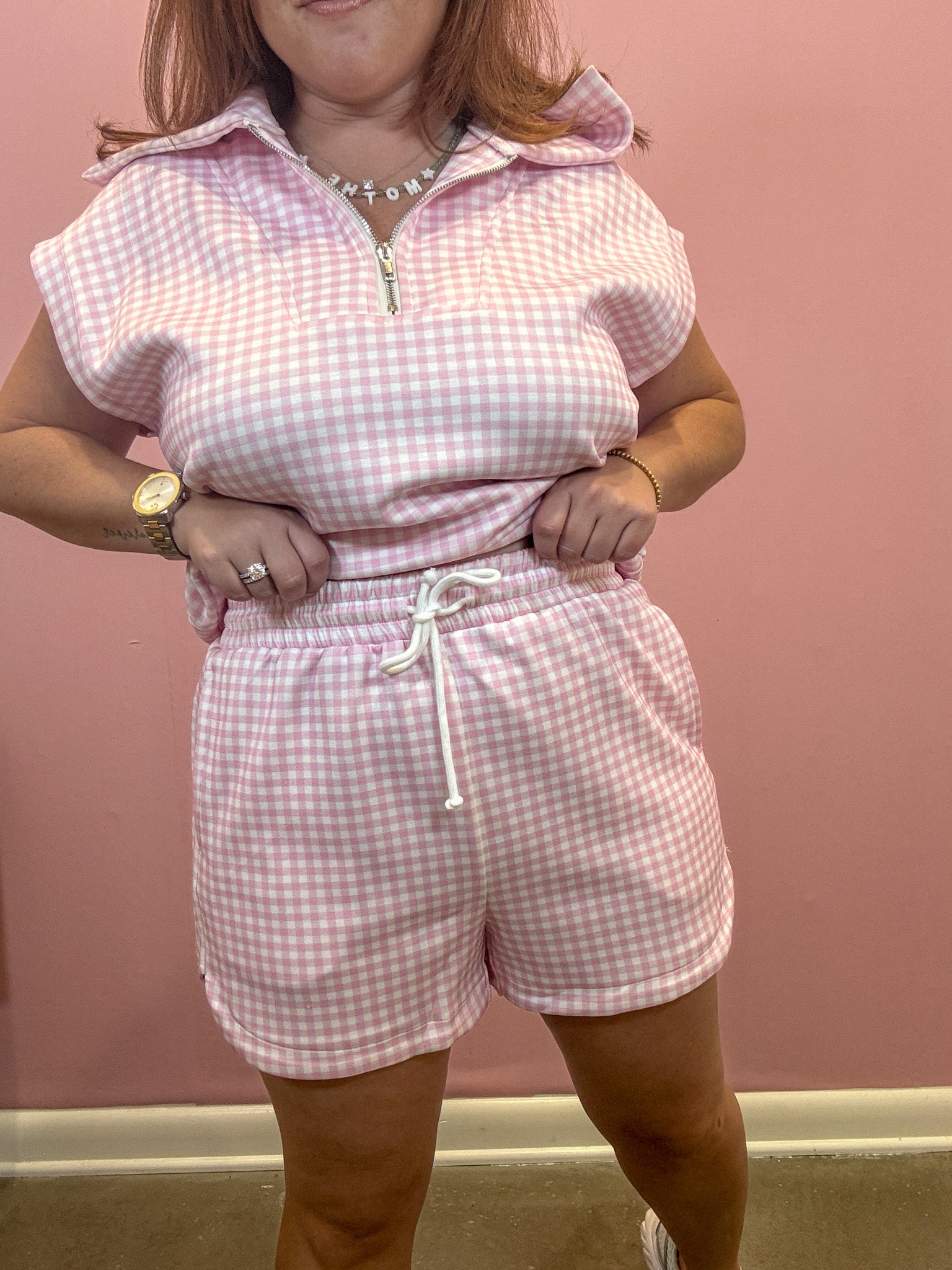 A woman modeling a pink and white gingham two-piece set consisting of a short-sleeve half-zip collared top and matching drawstring shorts, set against a solid pink wall.