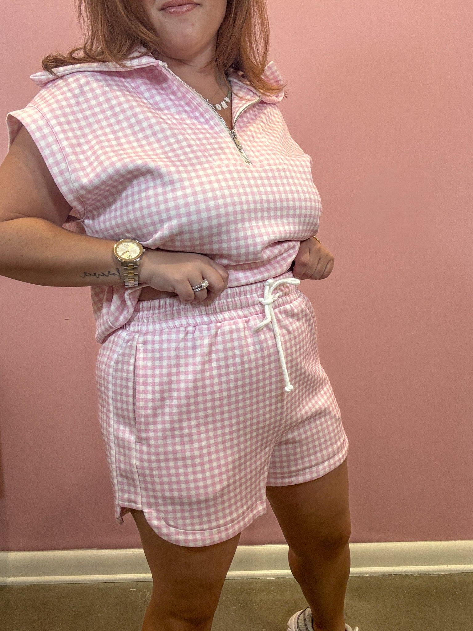 A woman modeling a pink and white gingham matching set consisting of a sleeveless half-zip collared top and matching drawstring shorts, standing against a solid pink wall.
