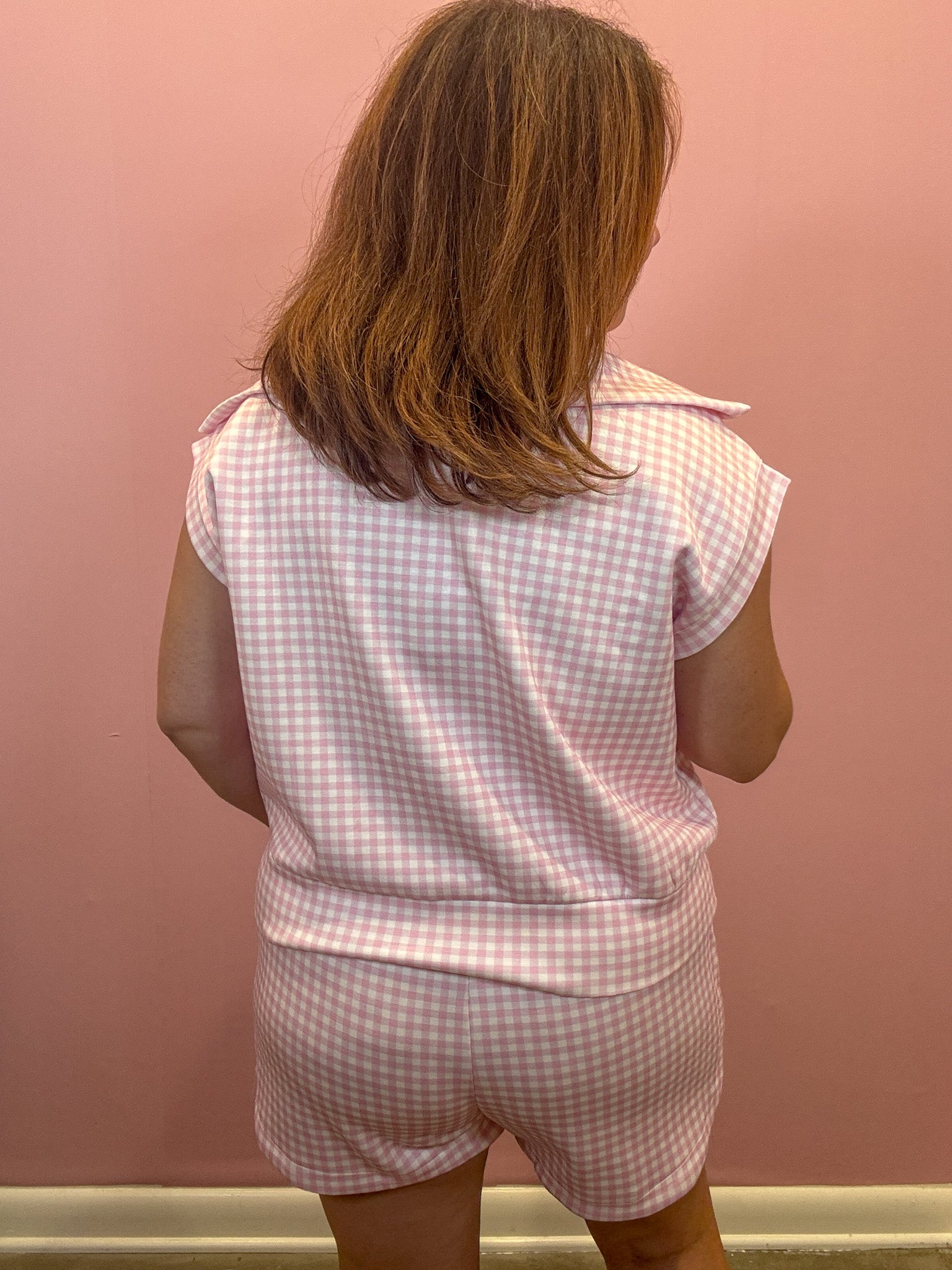 A child viewed from behind wearing a pink and white gingham short-sleeve top and matching shorts against a pink wall.