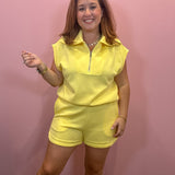 A woman modeling a bright yellow two-piece lounge set consisting of a sleeveless half-zip collared top and matching shorts, standing against a pink wall.