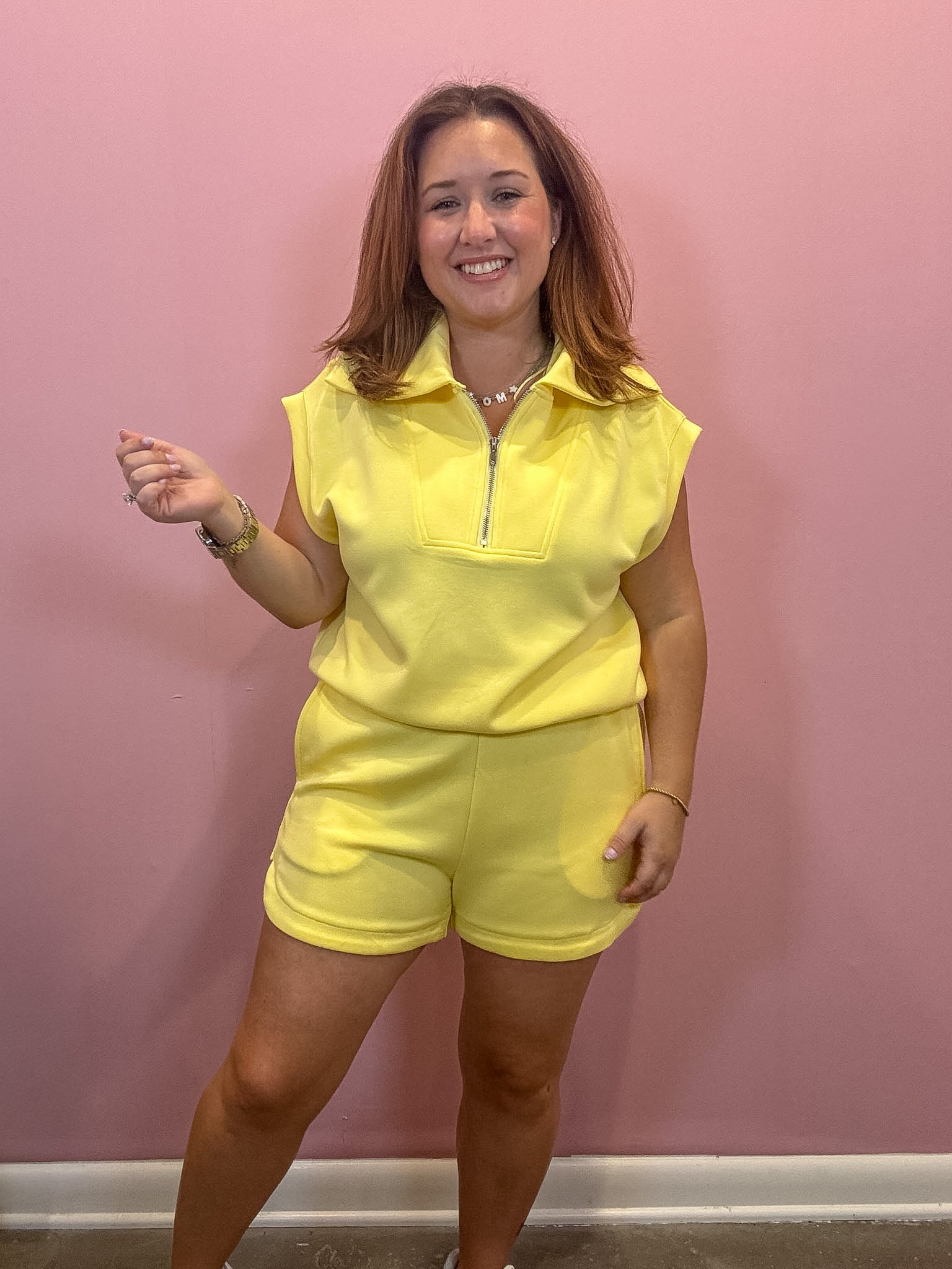 A woman modeling a bright yellow two-piece lounge set consisting of a sleeveless half-zip collared top and matching shorts, standing against a pink wall.