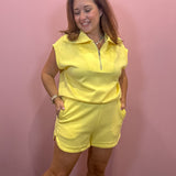 A woman modeling a bright yellow sleeveless zip-up polo shirt and matching shorts set against a solid pink wall.
