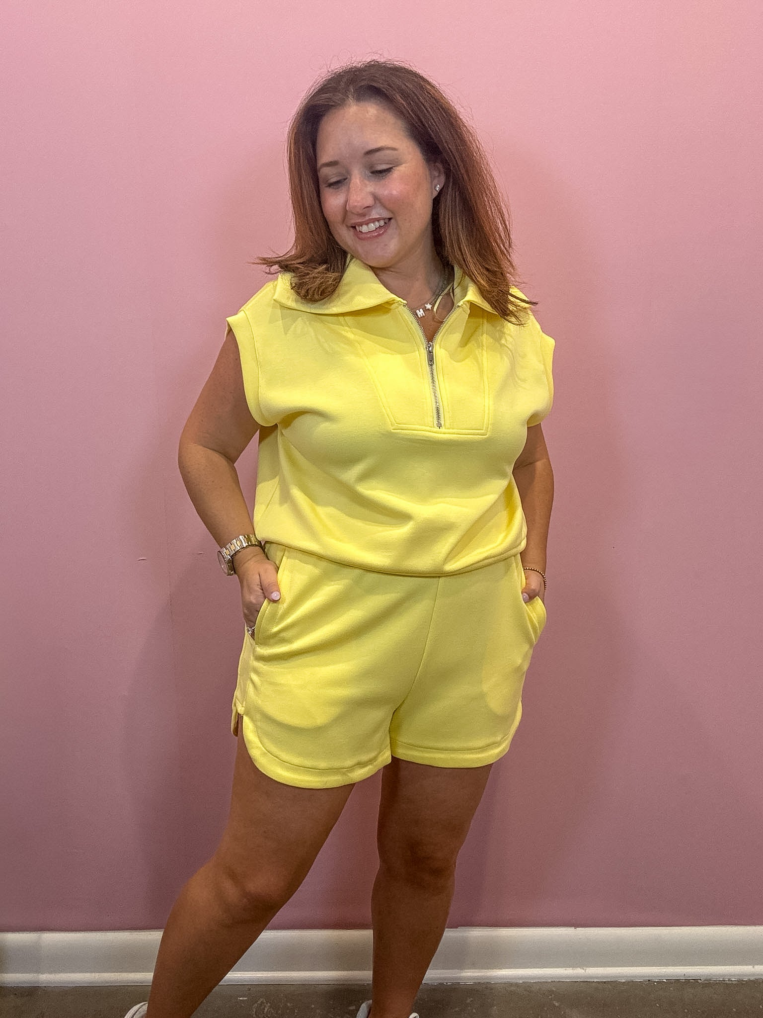 A woman modeling a bright yellow sleeveless zip-up polo shirt and matching shorts set against a solid pink wall.