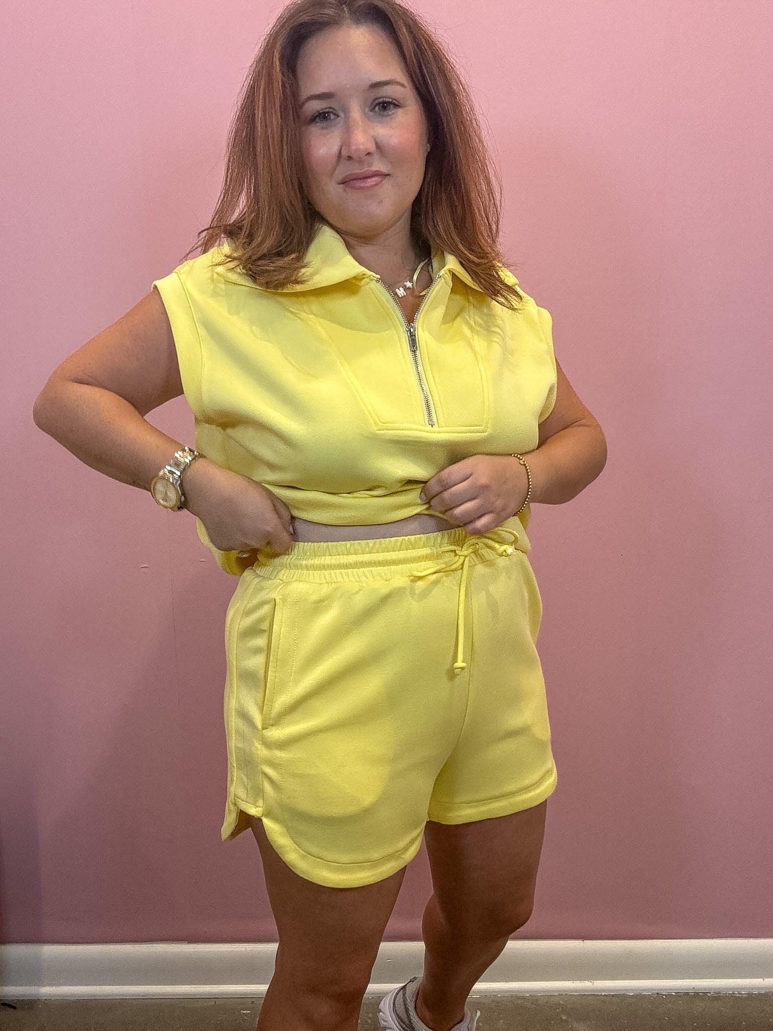 A woman modeling a bright yellow two-piece lounge set consisting of a sleeveless half-zip collared top and matching drawstring shorts, standing against a pink wall.