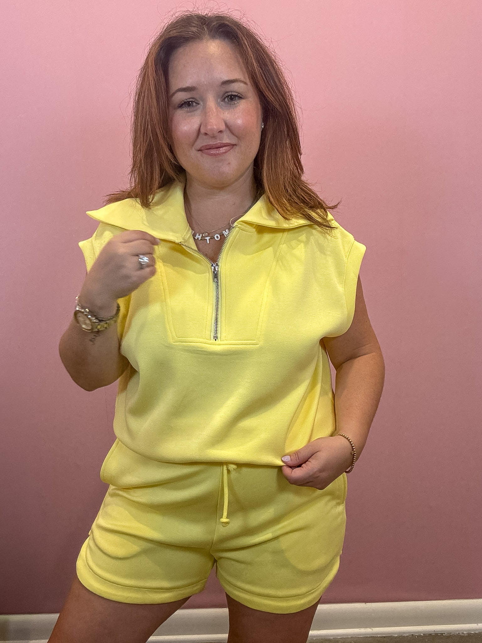 A woman modeling a bright yellow sleeveless zip-up top and matching shorts set against a solid pink wall.
