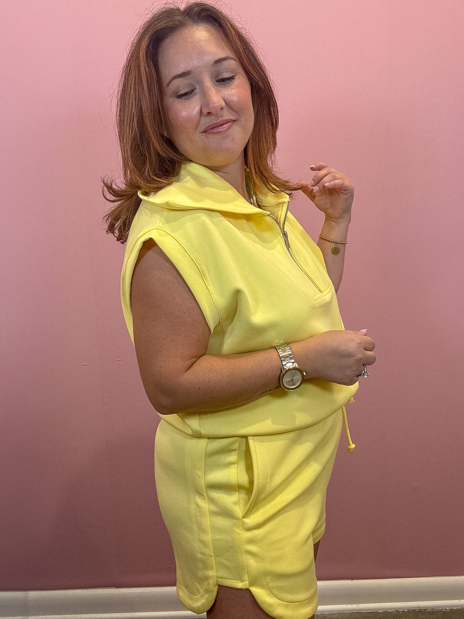 A woman wearing a bright yellow sleeveless zip-up hoodie and matching shorts set, standing against a solid pink wall.