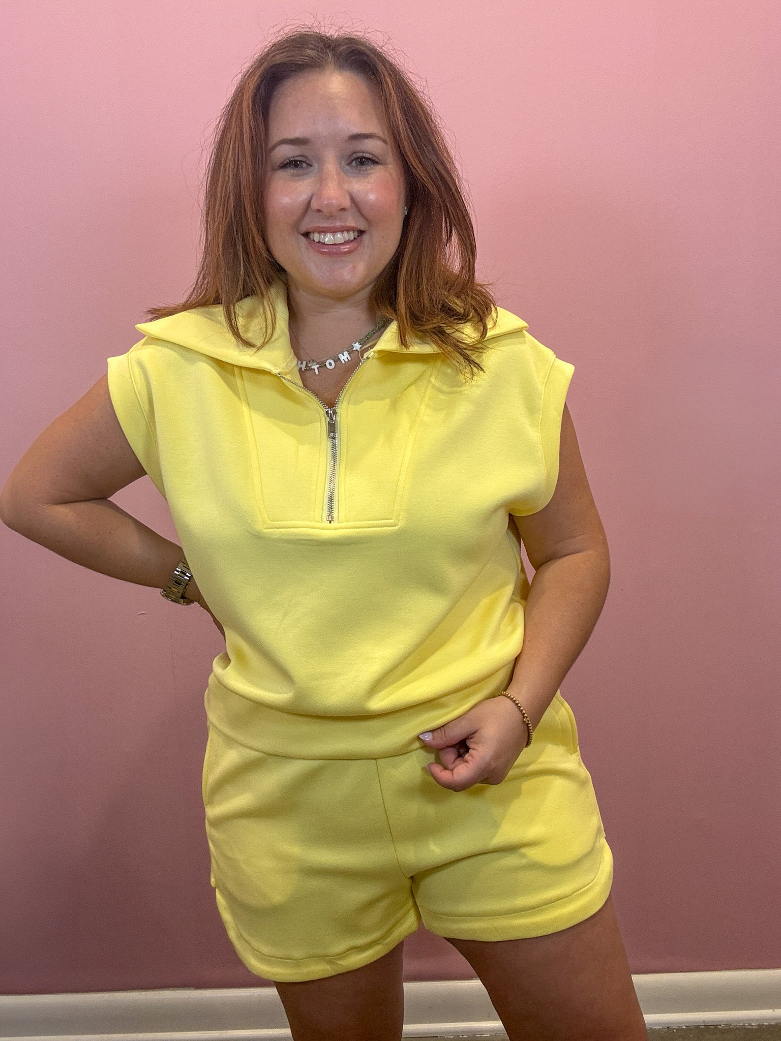 A woman modeling a bright yellow two-piece lounge set consisting of a short-sleeve half-zip pullover and matching shorts, standing against a solid pink wall.