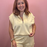 A woman modeling a cream-colored two-piece loungewear set consisting of a short-sleeve half-zip top and matching drawstring shorts, set against a solid pink wall.