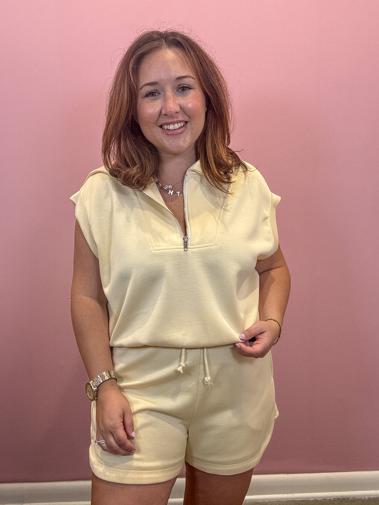 A woman modeling a cream-colored two-piece loungewear set consisting of a short-sleeve half-zip top and matching drawstring shorts, set against a solid pink wall.