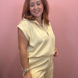 A woman modeling a pale yellow sleeveless quarter-zip pullover and matching shorts set against a solid pink wall.