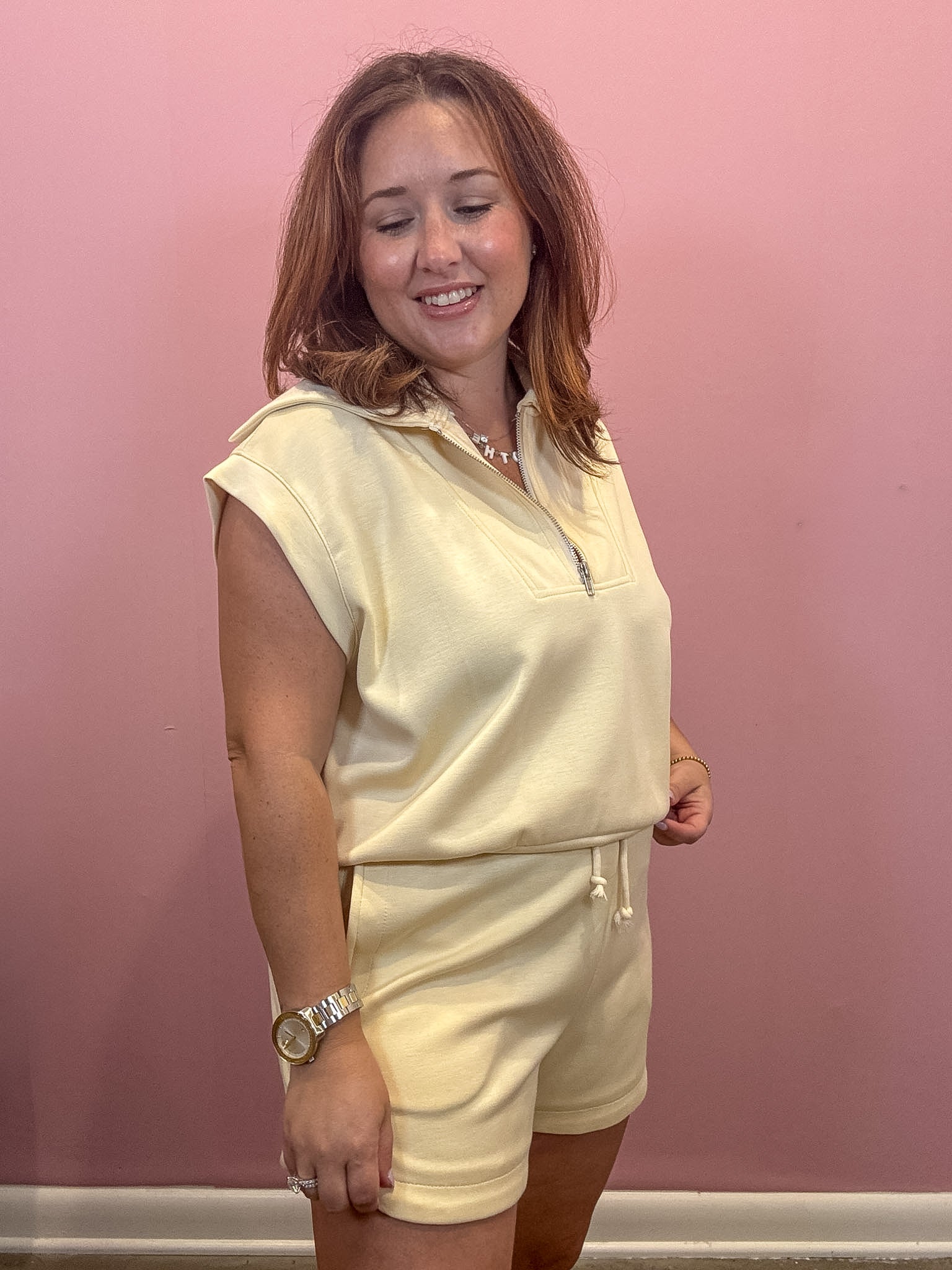 A woman modeling a pale yellow sleeveless quarter-zip pullover and matching shorts set against a solid pink wall.