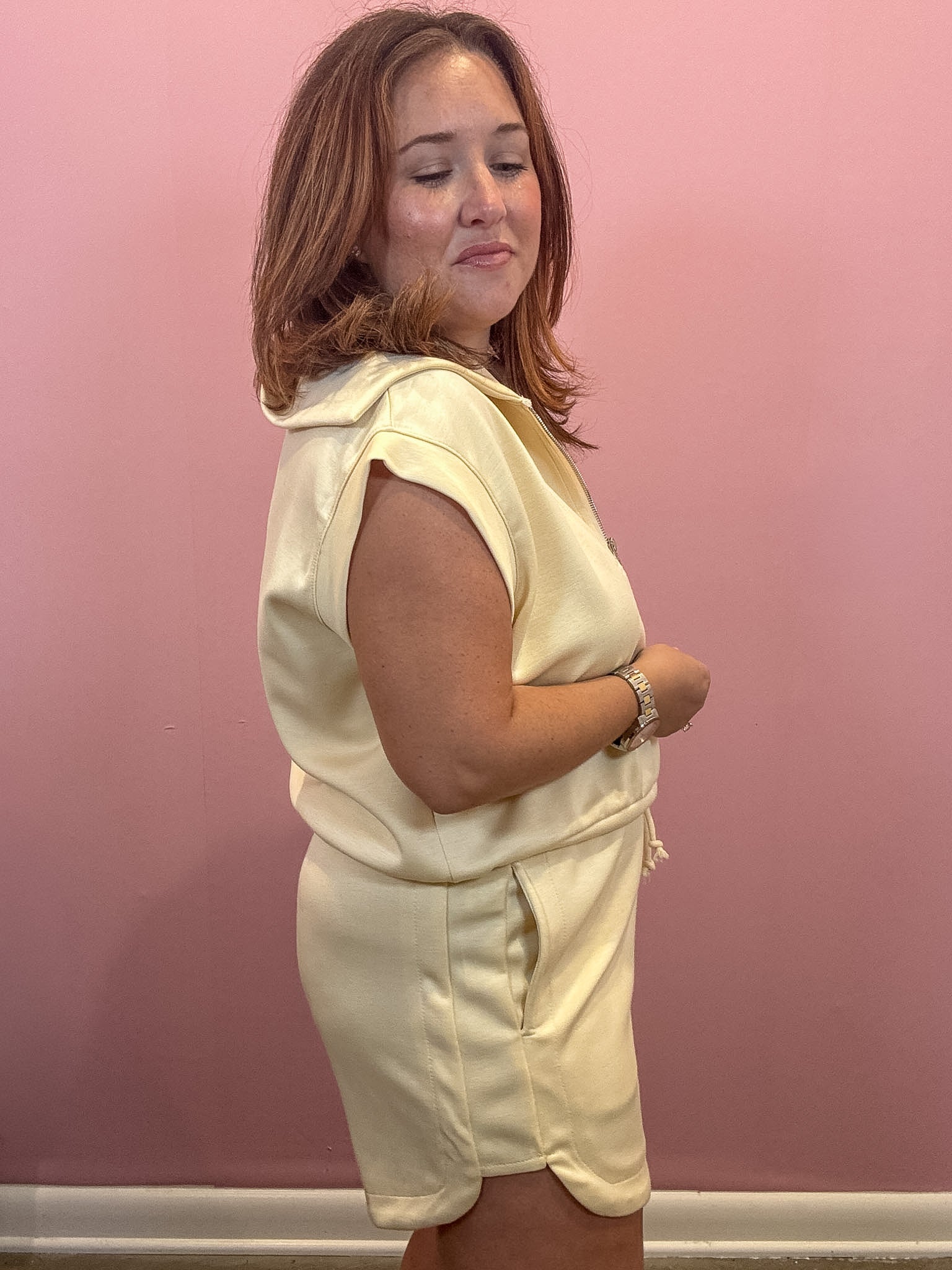 A woman wearing a cream-colored sleeveless hooded romper with side pockets, standing against a solid pink wall.