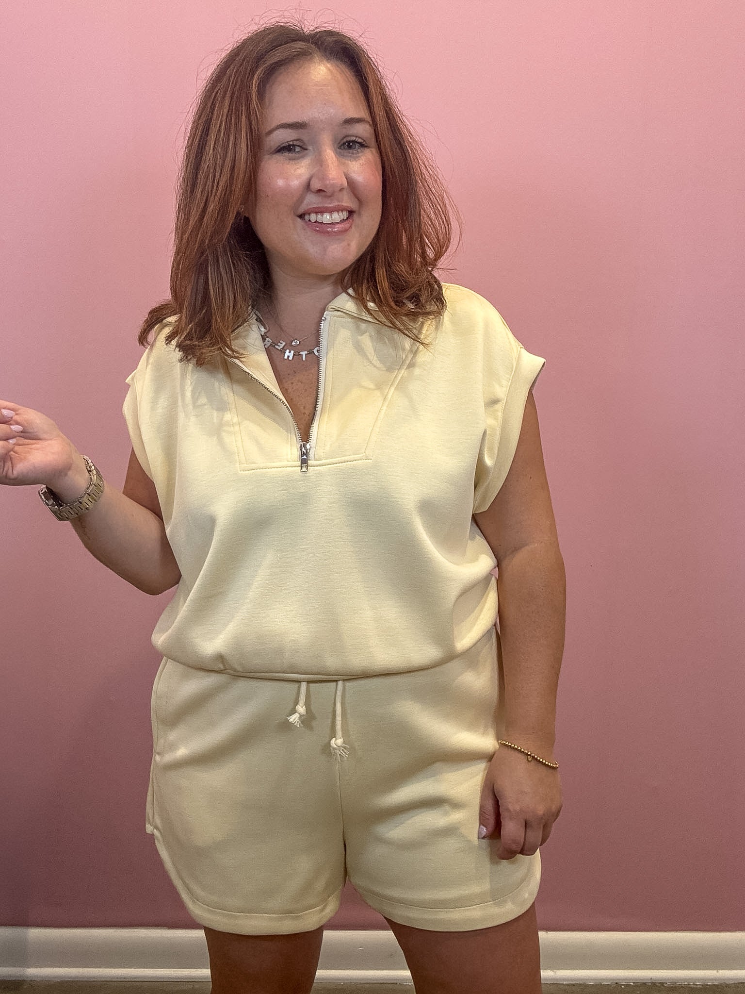 A woman modeling a cream-colored two-piece loungewear set consisting of a short-sleeve half-zip pullover and matching drawstring shorts, set against a solid pink wall.