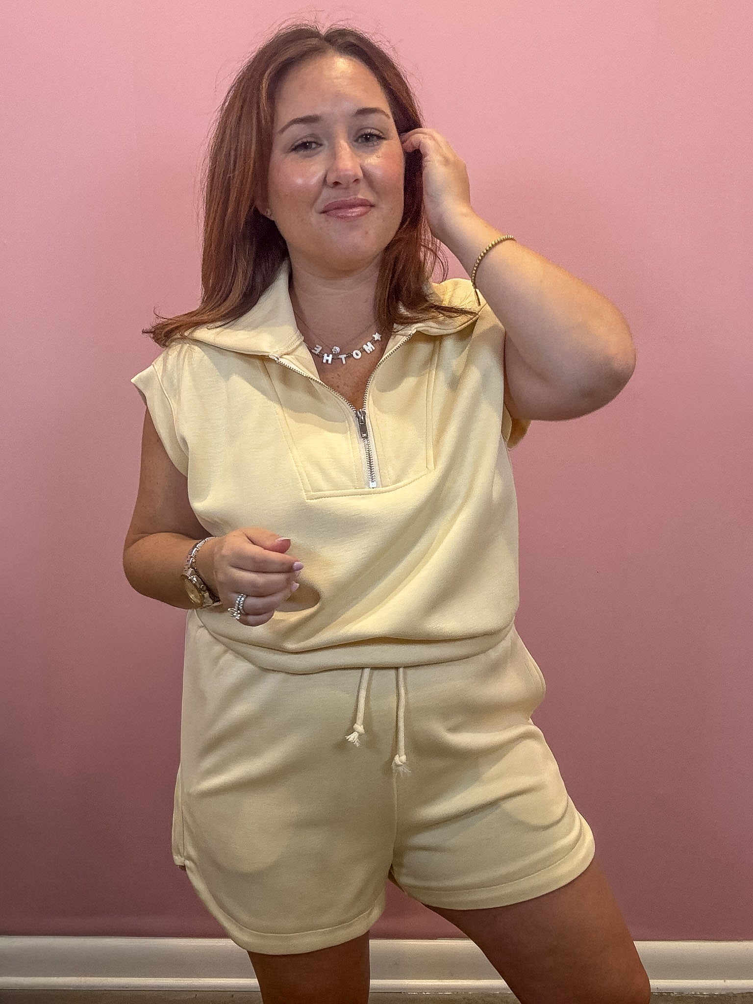 A woman modeling a pale yellow sleeveless collared pullover top and matching shorts set against a solid pink wall.