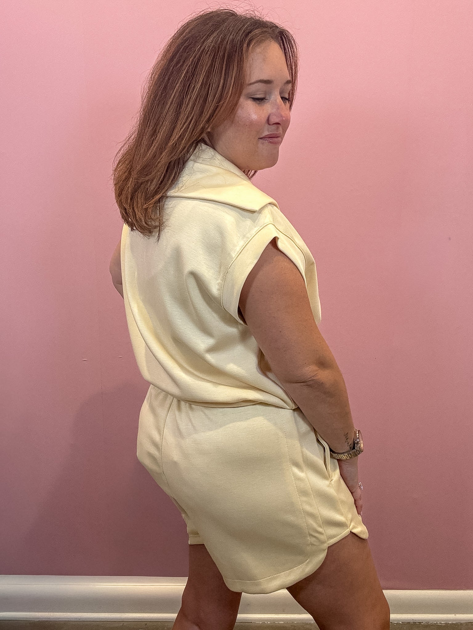 A woman wearing a pale yellow sleeveless collared top and matching shorts, standing against a pink wall.