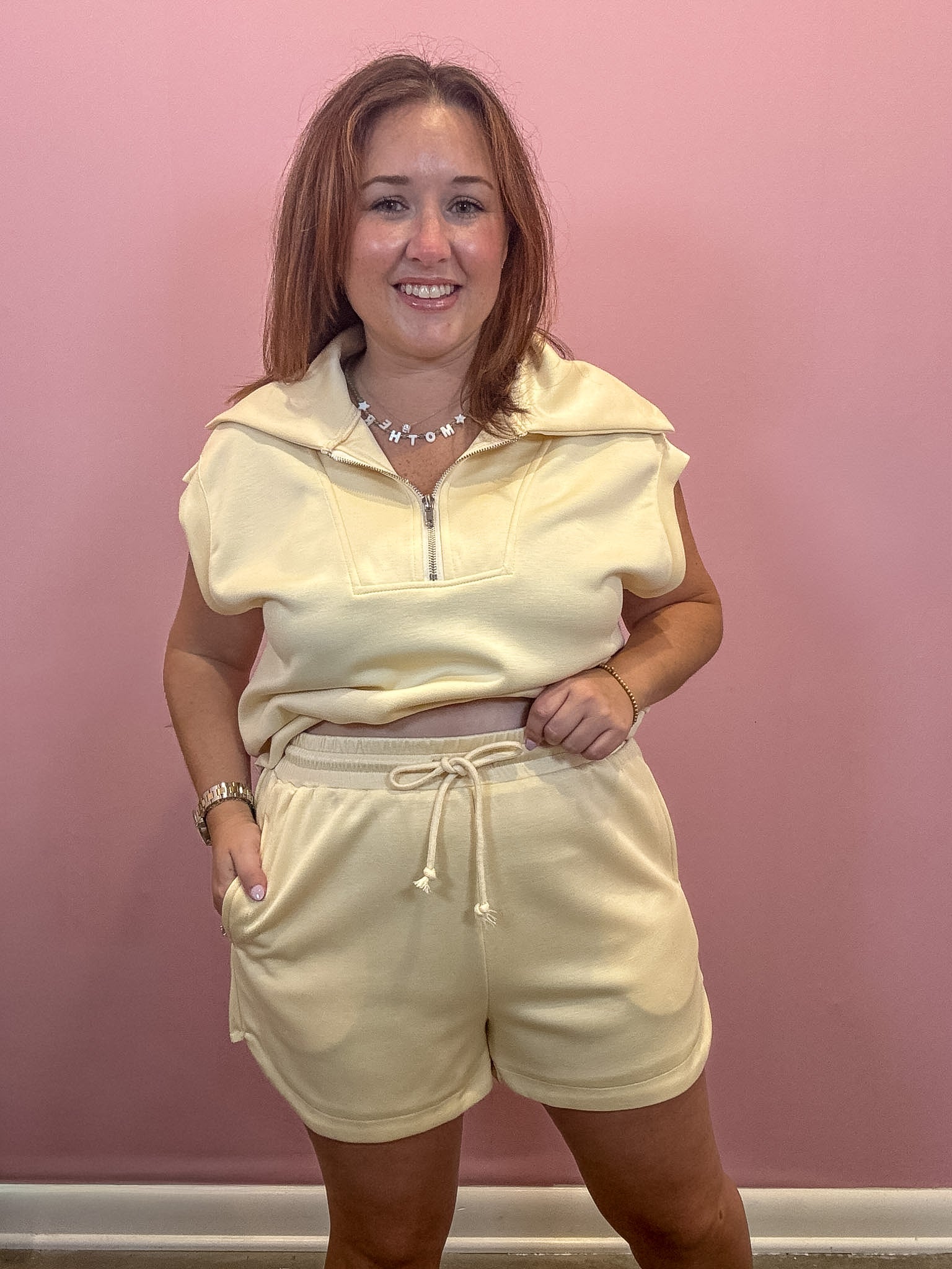 A woman modeling a pale yellow matching lounge set consisting of a short-sleeve half-zip pullover and drawstring shorts, standing against a pink wall.