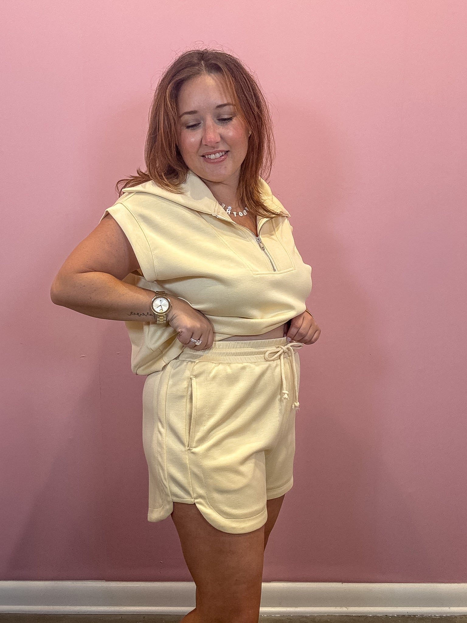 A woman modeling a pale yellow two-piece lounge set consisting of a sleeveless half-zip hoodie and matching shorts, standing against a solid pink wall.