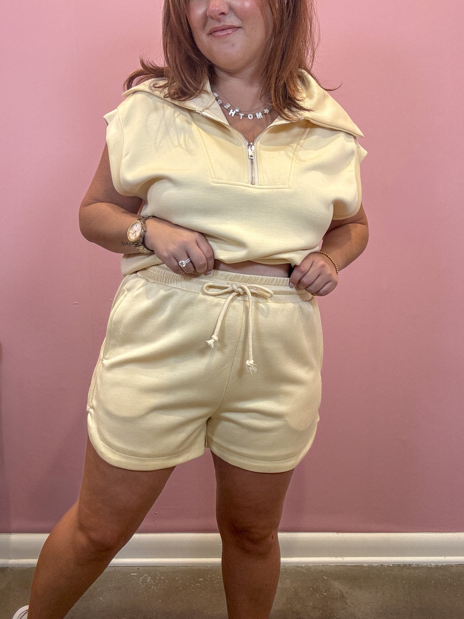 A woman modeling a matching pale yellow two-piece lounge set consisting of a short-sleeve half-zip pullover and drawstring shorts, posed against a solid pink wall.
