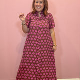 A woman modeling a short-sleeve button-up maxi dress with a pink and dark red block print pattern, standing against a solid pink wall.