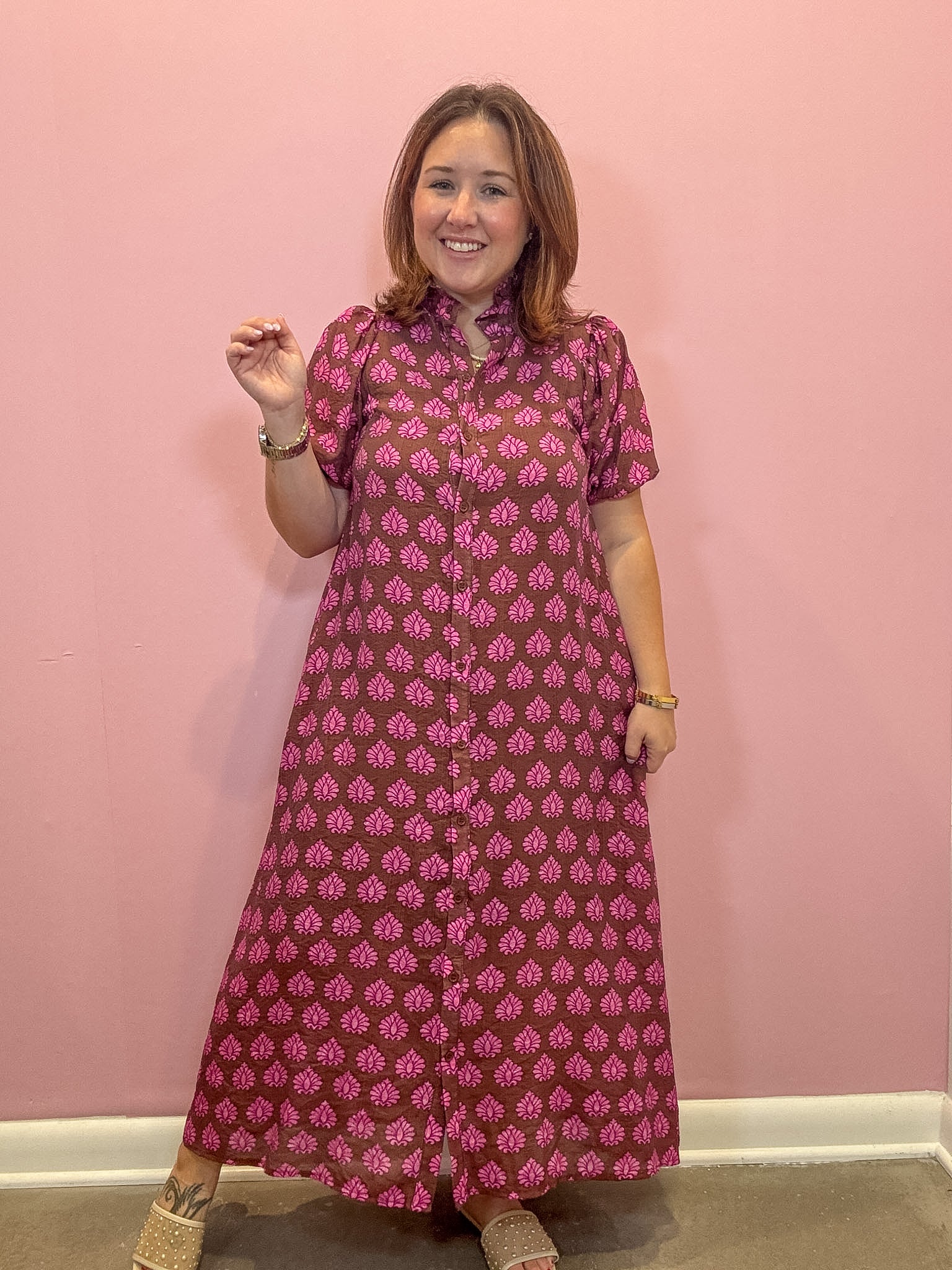 A woman modeling a short-sleeve button-up maxi dress with a pink and dark red block print pattern, standing against a solid pink wall.
